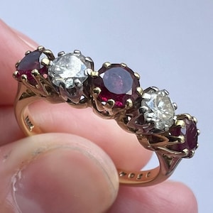 Puede incluir: Un anillo de oro con un engaste de cinco piedras que alterna rubíes de color rojo intenso y diamantes blancos transparentes de talla redonda. Las piedras están sujetas por garras de metal y la banda es de oro amarillo pulido con marcas visibles.