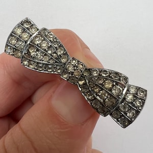 May include: A silver-toned bow-shaped brooch, encrusted with numerous small, clear rhinestones. The brooch is held in a hand against a white background, showcasing its intricate design and sparkling details. The bow design is a classic accessory.