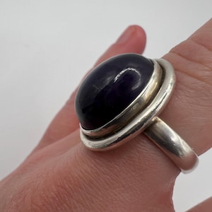 May include: A silver ring featuring a large, oval, dark purple gemstone. The gemstone is set in a bezel setting, and the ring has a simple, polished band. The ring is a statement piece, suitable for everyday wear or special occasions.