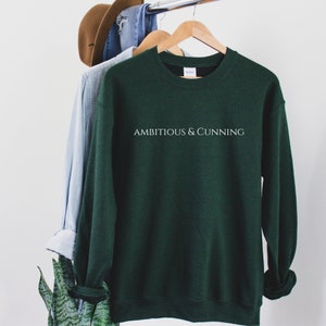May include: A dark green sweatshirt with the text "Ambitious & Cunning" printed in white on the front.
