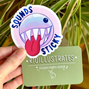 May include: A round sticker with a cartoon monster design. The monster has a blue and purple head, white teeth, and a red tongue. The sticker has the words "SOUNDS STICKY" in blue. A green card below reads "RIOILLUSTRATES".