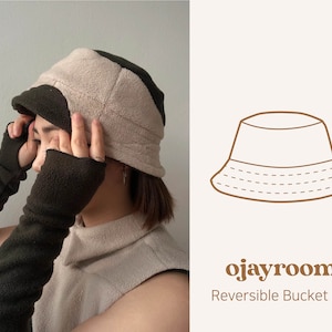 Pode incluir: Um chapéu de pescador reversível nas cores marrom e bege. O chapéu é feito de lã e tem uma costura em volta da aba. O texto "ojayroom Reversible Bucket Hat" está impresso abaixo do chapéu.