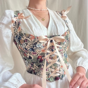 K&ouml;nnte beinhalten: Ein cremefarbenes Korsett mit einem floralen Tapisserie-Muster und einer Schn&uuml;rung vorne. Das Korsett hat ein Satinband, das an den Schultern gebunden wird, und ein passendes Band, das vorne geschn&uuml;rt wird. Das Korsett wird &uuml;ber einem wei&szlig;en Oberteil getragen.