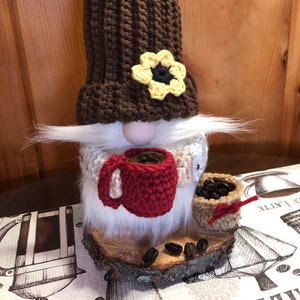 May include: A brown crocheted gnome wearing a yellow flower on its hat, holding a red crocheted coffee mug and a small brown bag of coffee beans. The gnome is standing on a wooden slice with coffee beans scattered around it.