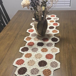 May include: A crocheted table runner with a hexagonal pattern in shades of brown, beige, and orange. The runner is made of yarn and has a textured surface.