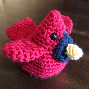 May include: A crocheted cardinal bird toy with red, black, and yellow yarn. The bird has a small beak and black eyes.