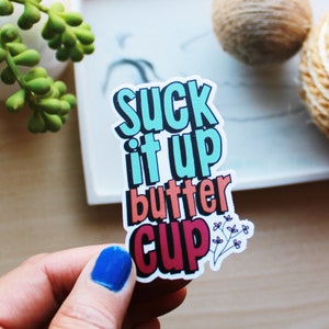 May include: A sticker with the text "Suck it up butter cup" in colorful, bold letters. The sticker has a white background and is held by a hand with blue nail polish.