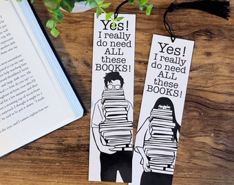 May include: Two white bookmarks with black illustrations of people holding stacks of books. The text on the bookmarks reads "Yes! I really do need ALL these BOOKS!"