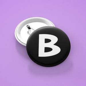May include: A black button with a white letter "B" on it. The button is attached to a silver pin.