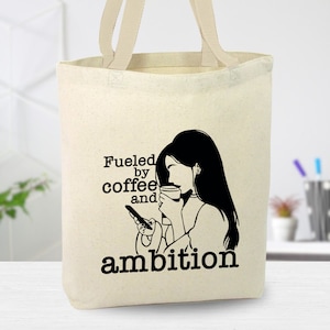 May include: A white canvas tote bag with a black silhouette of a woman drinking coffee and looking at her phone. The text "Fueled by coffee and ambition" is printed on the bag.