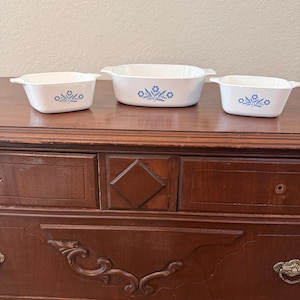 Könnte beinhalten: Drei weiße Auflaufformen mit blauen Blumenmustern sind auf einer Holzoberfläche angeordnet. Jede Form hat eine rechteckige Form mit abgerundeten Ecken und Griffen. Das blaue Blumenmuster ist auf jeder Form zentriert.