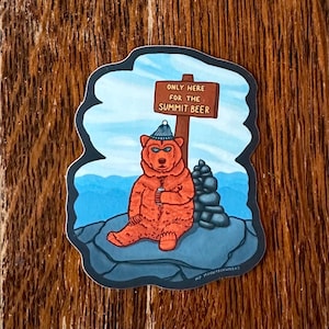 May include: A sticker featuring a cartoon bear wearing a blue beanie, holding a can, and sitting on a rock. A sign reads "ONLY HERE FOR THE SUMMIT BEER." The background shows a mountain scene with blue sky and water.