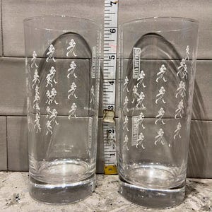 May include: Two clear highball glasses with a repeating white graphic of a striding man. The glasses are approximately 6 inches tall. The text "KEEPS MY KING" is printed on the side of each glass.