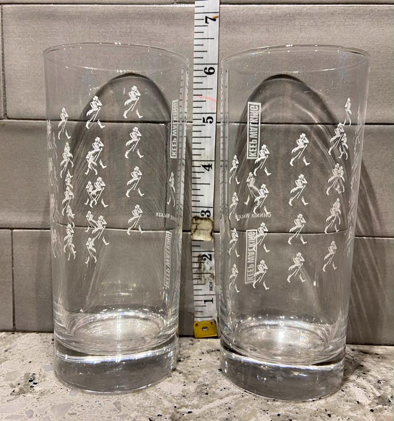 May include: Two clear highball glasses with a repeating white graphic of a striding man. The glasses are approximately 6 inches tall. The text "KEEPS MY KING" is printed on the side of each glass.