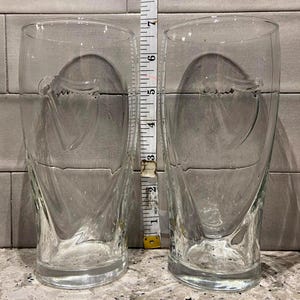 May include: Two clear, tall drinking glasses with a decorative swirl design. Each glass is approximately 6 inches tall. The glasses are made of clear glass and are empty. A measuring tape is placed between the glasses.