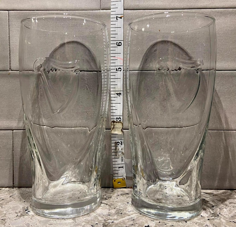 May include: Two clear, tall drinking glasses with a decorative swirl design. Each glass is approximately 6 inches tall. The glasses are made of clear glass and are empty. A measuring tape is placed between the glasses.