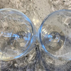 May include: Two clear stemmed wine glasses, viewed from above, are positioned next to each other. The glasses are empty and made of glass. The background is a speckled, light-coloured surface.