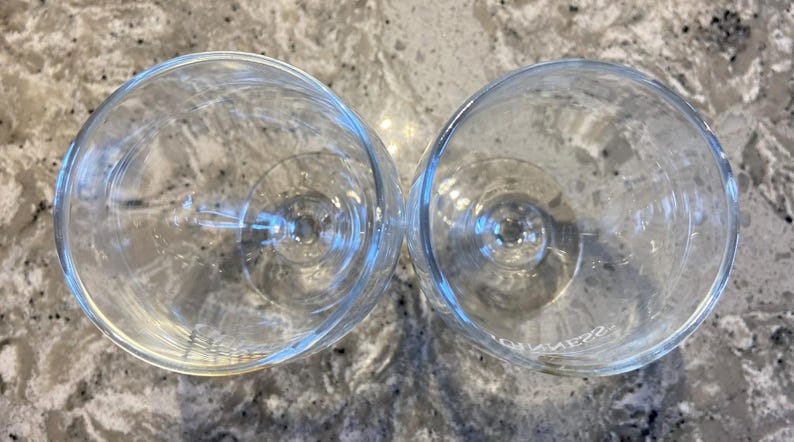 May include: Two clear stemmed wine glasses, viewed from above, are positioned next to each other. The glasses are empty and made of glass. The background is a speckled, light-coloured surface.
