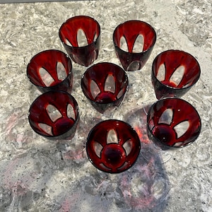 Op de afbeelding: Set van acht rode glazen wijnglazen met een helder, uitgesneden ontwerp. De glazen zijn in een cirkel op een lichtgrijs oppervlak gerangschikt.