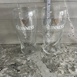 K&ouml;nnte beinhalten: Zwei transparente Guinness-Biergl&auml;ser mit dem Guinness-Logo und -Text. Ein Ma&szlig;band zeigt, dass die Gl&auml;ser etwa 15 cm hoch sind. Die Gl&auml;ser stehen auf einer gesprenkelten grauen Arbeitsplatte.