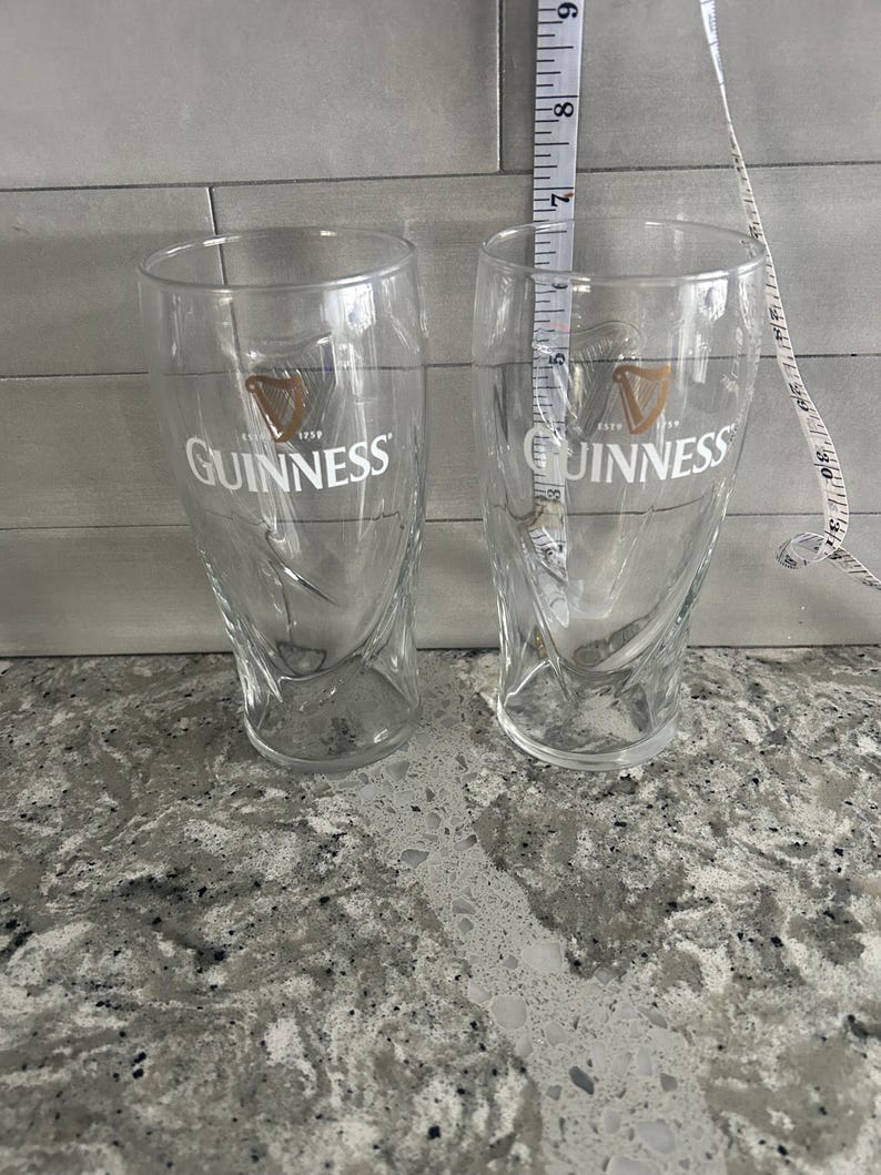 K&ouml;nnte beinhalten: Zwei transparente Guinness-Biergl&auml;ser mit dem Guinness-Logo und -Text. Ein Ma&szlig;band zeigt, dass die Gl&auml;ser etwa 15 cm hoch sind. Die Gl&auml;ser stehen auf einer gesprenkelten grauen Arbeitsplatte.