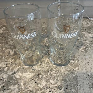 K&ouml;nnte beinhalten: Zwei transparente Guinness-Biergl&auml;ser mit dem Guinness-Logo in Wei&szlig; und Gold. Die Gl&auml;ser haben ein einzigartiges, geschwungenes Design und stehen auf einer gesprenkelten Arbeitsplatte.