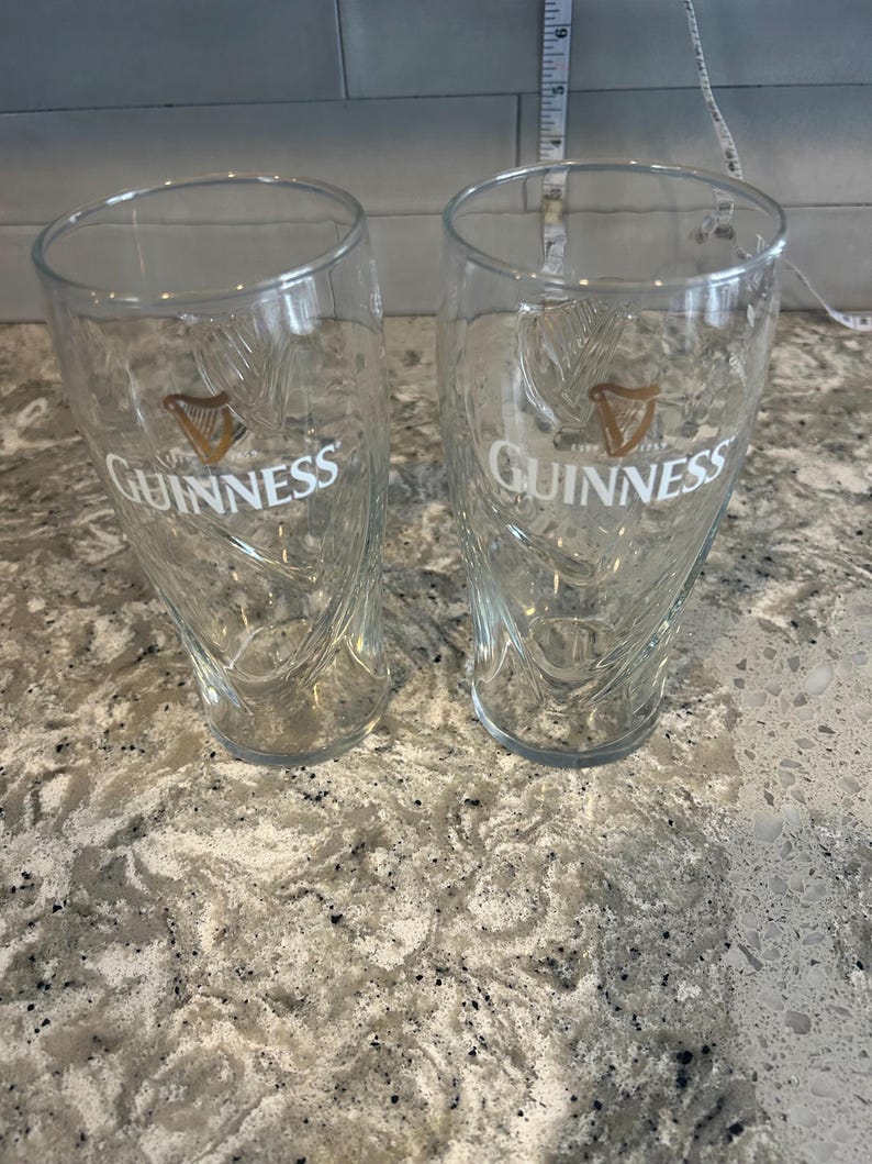 K&ouml;nnte beinhalten: Zwei transparente Guinness-Biergl&auml;ser mit dem Guinness-Logo in Wei&szlig; und Gold. Die Gl&auml;ser haben ein einzigartiges, geschwungenes Design und stehen auf einer gesprenkelten Arbeitsplatte.