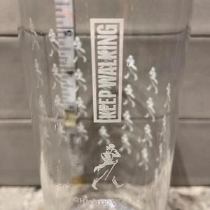 May include: Clear pint glass with the text "KEEP WALKING" in a white rectangular box. The glass features a white silhouette of a man walking with a cane, and small white figures walking around the glass. The words "JOHNNIE WALKER" are printed at the bottom.