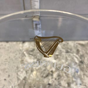 May include: A clear glass with the Guinness logo, featuring a gold harp emblem and the word "Guinness" in white lettering. A measurement tool is visible in the background.