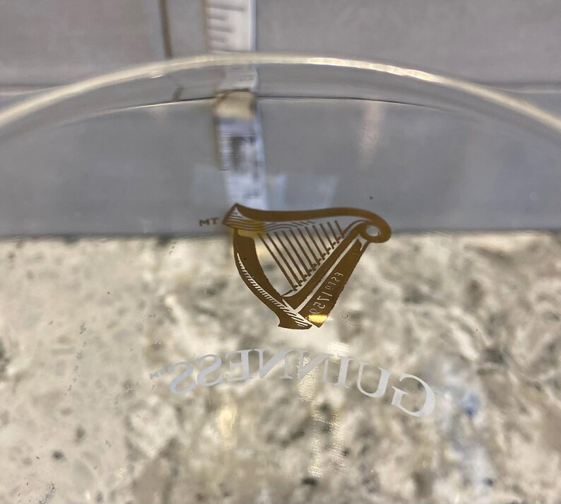May include: A clear glass with the Guinness logo, featuring a gold harp emblem and the word "Guinness" in white lettering. A measurement tool is visible in the background.