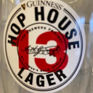 May include: Clear glass with a circular Guinness Hop House 13 Lager sticker. The sticker features the Guinness logo, the words "HOP HOUSE" and "LAGER", and a large red "13". The text "The Brewers Project" and "St. James's Gate, Dublin" are also visible.