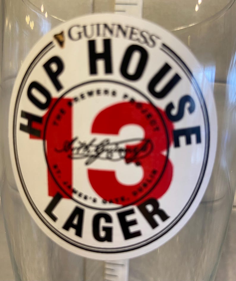May include: Clear glass with a circular Guinness Hop House 13 Lager sticker. The sticker features the Guinness logo, the words "HOP HOUSE" and "LAGER", and a large red "13". The text "The Brewers Project" and "St. James's Gate, Dublin" are also visible.
