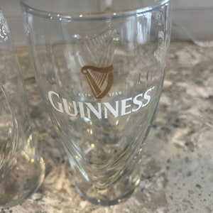 K&ouml;nnte beinhalten: Klares Guinness-Bierglas mit dem Guinness-Logo in Gold und Wei&szlig;. Das Glas hat ein einzigartiges, geschwungenes Design und der Text "ESTD 1759" ist darunter zu sehen.
