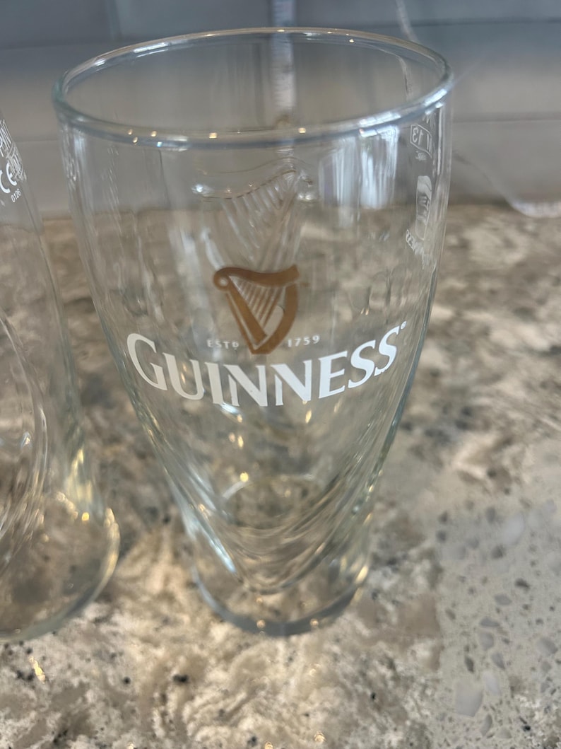K&ouml;nnte beinhalten: Klares Guinness-Bierglas mit dem Guinness-Logo in Gold und Wei&szlig;. Das Glas hat ein einzigartiges, geschwungenes Design und der Text "ESTD 1759" ist darunter zu sehen.