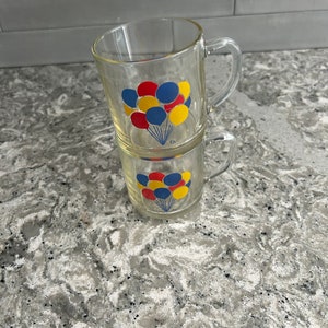 Puede incluir: Dos tazas de vidrio transparente con asas, apiladas una encima de la otra. Las tazas tienen un diseño de globos rojos, azules y amarillos con cuerdas.