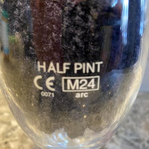 May include: A clear glass with the words "HALF PINT" in white. Below the text are the letters "CE" and the number "0071". A box with "M24" and the word "arc" is also visible. The glass is partially filled with a dark liquid.