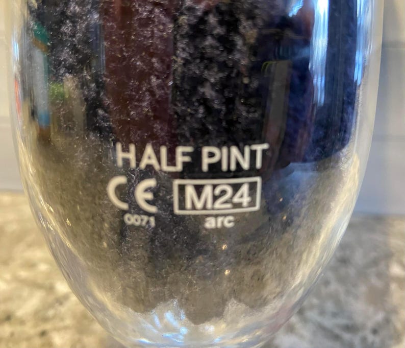May include: A clear glass with the words "HALF PINT" in white. Below the text are the letters "CE" and the number "0071". A box with "M24" and the word "arc" is also visible. The glass is partially filled with a dark liquid.