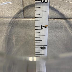 May include: A clear glass container with a white measuring tape inside. The tape shows a measurement of approximately 7.5 inches. The container is on a light gray tiled surface.