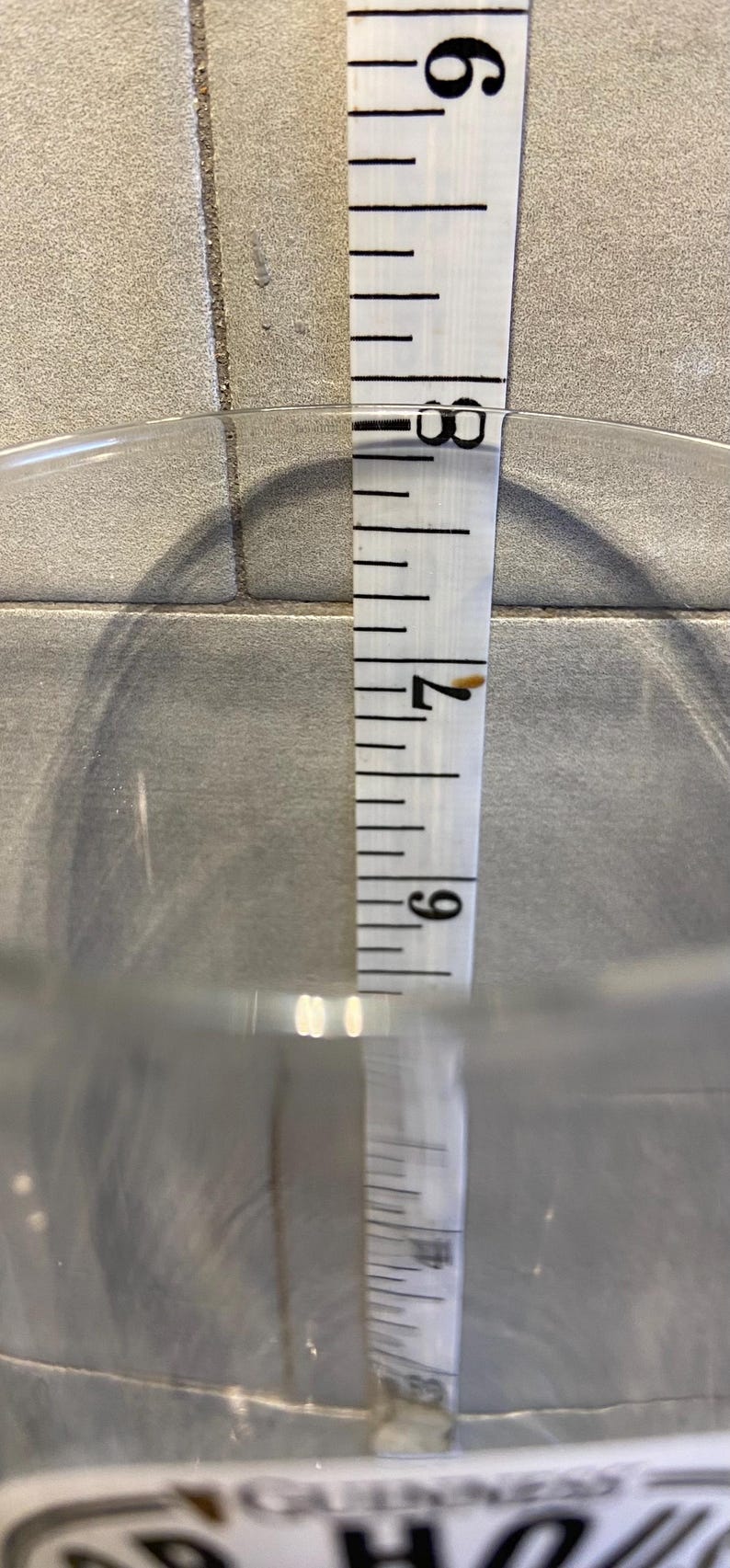 May include: A clear glass container with a white measuring tape inside. The tape shows a measurement of approximately 7.5 inches. The container is on a light gray tiled surface.