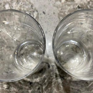May include: Two clear, empty drinking glasses. The glasses are cylindrical with a rounded base and are made of transparent glass. The glasses are positioned side by side on a speckled, light-colored surface.