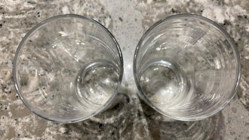 May include: Two clear, empty drinking glasses. The glasses are cylindrical with a rounded base and are made of transparent glass. The glasses are positioned side by side on a speckled, light-colored surface.