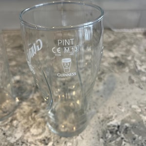 K&ouml;nnte beinhalten: Ein klares Pint-Glas mit dem Guinness-Logo und Text. Das Glas hat eine gebogene Form und eine breite &Ouml;ffnung. Der Text "PINT" und "2.3 UK UNITS" sind sichtbar. Das Glas ist leer. Es fasst 568 ml.