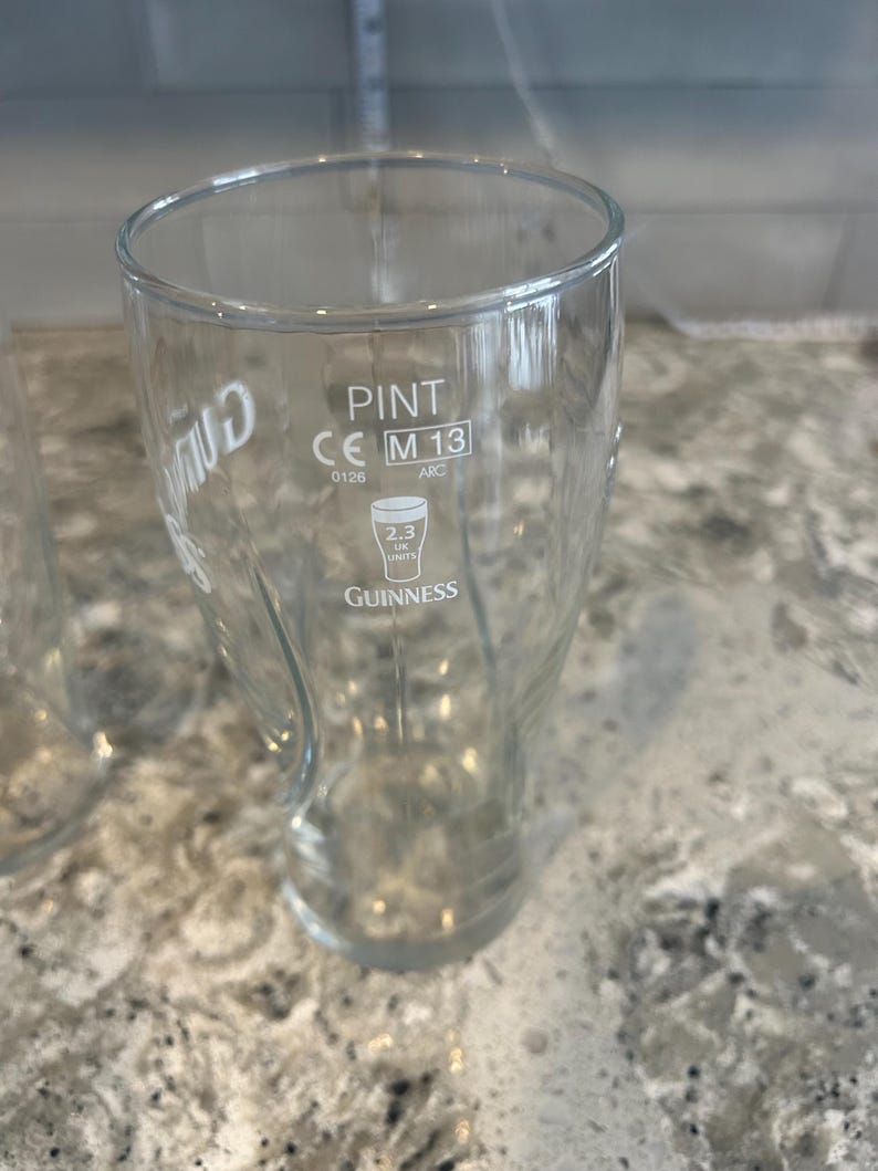 K&ouml;nnte beinhalten: Ein klares Pint-Glas mit dem Guinness-Logo und Text. Das Glas hat eine gebogene Form und eine breite &Ouml;ffnung. Der Text "PINT" und "2.3 UK UNITS" sind sichtbar. Das Glas ist leer. Es fasst 568 ml.