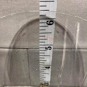 May include: A clear glass bowl with a diameter of approximately 6 inches is measured with a white measuring tape. The tape shows the bowl's depth. The bowl is on a gray surface.