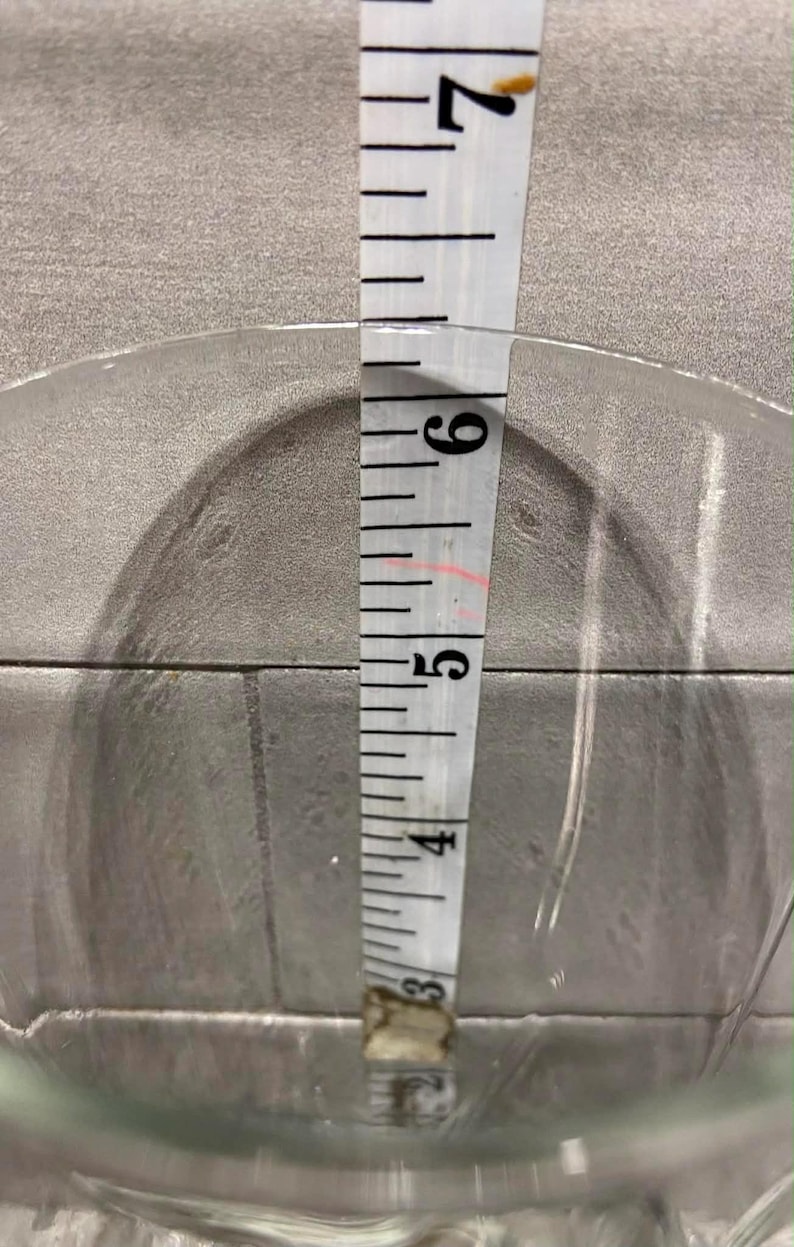 May include: A clear glass bowl with a diameter of approximately 6 inches is measured with a white measuring tape. The tape shows the bowl's depth. The bowl is on a gray surface.