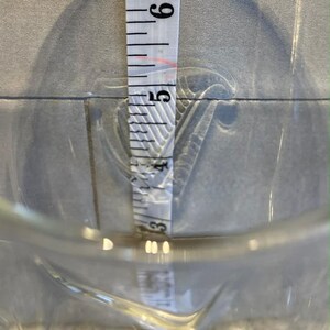 May include: A clear glass measuring cup with a pouring spout. A white measuring tape is inside the cup, showing a measurement of approximately 5 inches. The background is a light gray tile.