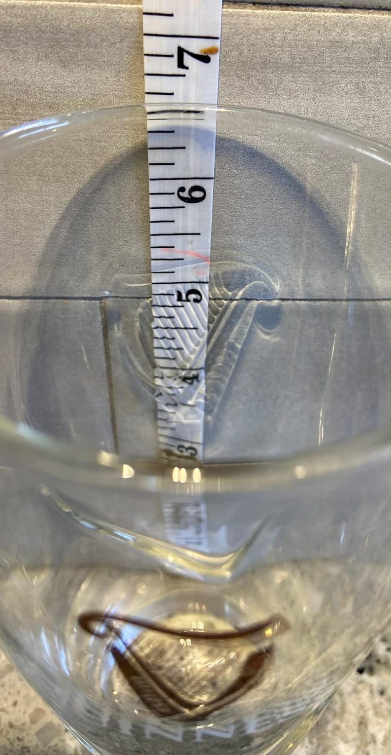 May include: A clear glass measuring cup with a pouring spout. A white measuring tape is inside the cup, showing a measurement of approximately 5 inches. The background is a light gray tile.