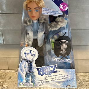 Könnte beinhalten: Bratz Boyz Wintertime Cameron Puppe in einer transparenten Kunststoffverpackung. Die Puppe hat blondes Haar, blaue Augen und trägt eine silberne Jacke mit Kunstfellbesatz. Zubehör umfasst einen Rucksack und einen Schal. Der Text "Wintertime Collection".