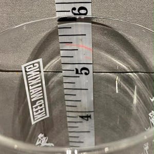 May include: A clear glass with a measurement of approximately 5 inches, as indicated by a white measuring tape. The glass has a sticker with the text "KEEB MYTKMC".
