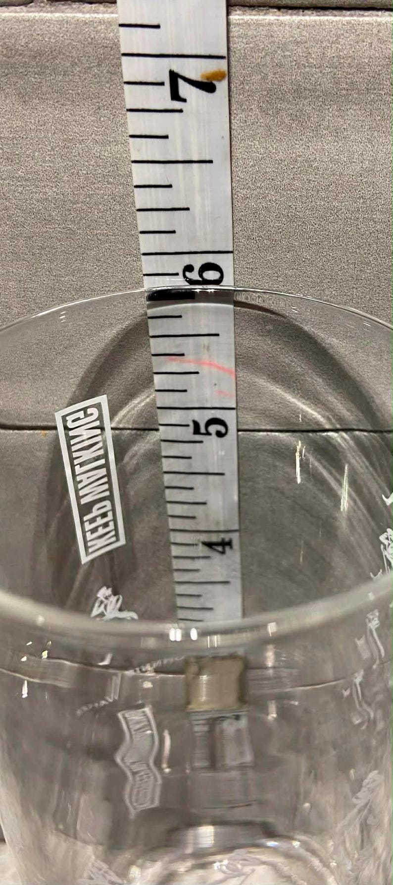 May include: A clear glass with a measurement of approximately 5 inches, as indicated by a white measuring tape. The glass has a sticker with the text "KEEB MYTKMC".